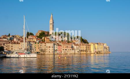 Mentre il sole tramonta su Rovigno, il cielo è dipinto in tonalità calde, gettando un bagliore dorato sul pittoresco lungomare. Edifici colorati fiancheggiano la riva, accompagnati da tranquille barche a vela nel porto. Foto Stock