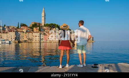 Una coppia si trova mano nella mano sul lungomare, ammirando la pittoresca Rovigno, Croazia. Gli edifici vibranti e la torreggiante torre dell'orologio si riflettono splendidamente nelle acque calme sotto un cielo azzurro. Foto Stock