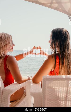 Due donne in costumi da bagno rossi fanno un cuore con le mani su una spiaggia Foto Stock