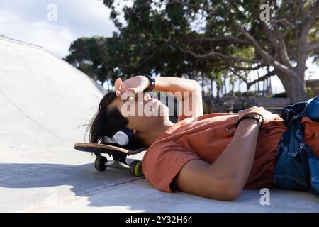 Riposante sullo skateboard, adolescente asiatico che ascolta musica con le cuffie all'aperto Foto Stock