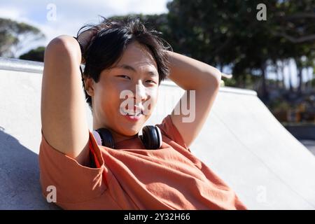 All'aperto, rilassatevi allo skatepark, adolescente asiatico con bretelle e cuffie sorridenti Foto Stock