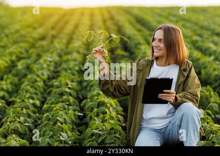 Tecnologia intelligente della soia per l'agricoltura. Una donna agricoltrice con tablet digitale esamina e controlla le radici delle piante di soia nel campo. L'agronomo controlla la crescita e lo sviluppo dei germogli prima della raccolta Foto Stock