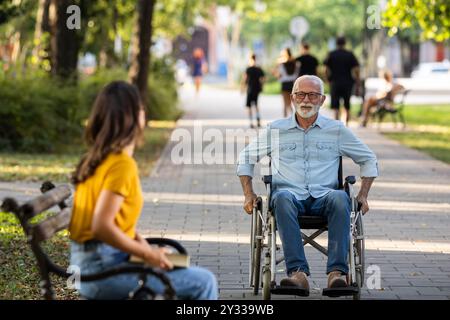 Uomo anziano che usa la sedia a rotelle mentre la giovane donna seduta sulla panchina e aspetta nel parco Foto Stock