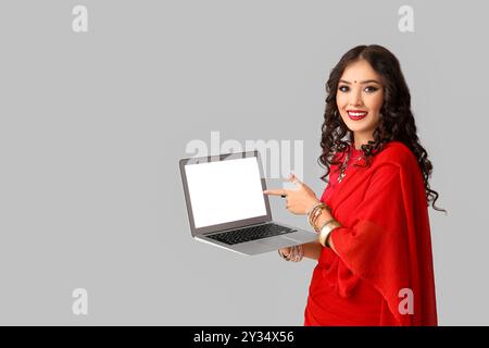 Bella donna indiana in sari che punta il laptop su sfondo grigio Foto Stock