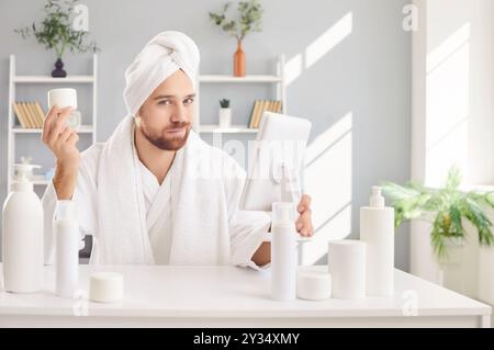 Uomo divertente in accappatoio dopo la doccia che finge di essere bellissimo, idratante sulla pelle, guardando allo specchio Foto Stock