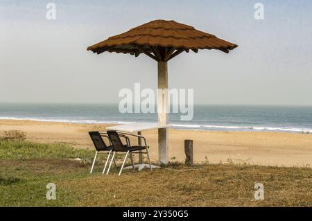 Due sedie e un tavolo sono posti di fronte al mare, appena sotto un ombrello di piastrelle rosse su una spiaggia in Gambia Foto Stock