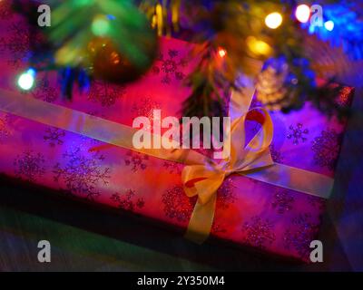 Un regalo di Natale in un involucro rosso si trova sotto un albero di Natale decorato con luci colorate. Concetto di celebrazione di Natale e Capodanno. Sfondo W Foto Stock