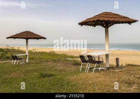 Due sedie e un tavolo sono posti di fronte al mare, appena sotto un ombrello di piastrelle rosse su una spiaggia in Gambia Foto Stock