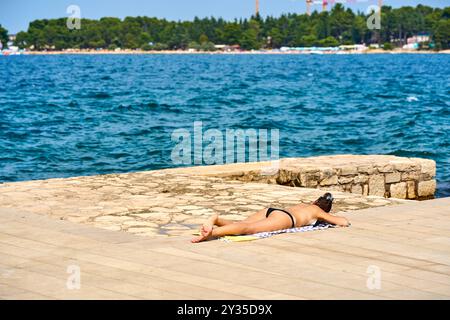 Porec, Istria, Croazia - 27 agosto 2024: Una donna si prende il sole su una piattaforma di pietra in riva al mare e gode dell'estate *** Eine Frau sonnt sich auf einer Steinplattform am Meer und genießt den Sommer Foto Stock