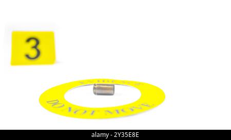 Un involucro di proiettile esaurito collocato all'interno di un cerchio giallo, etichettato con il testo "Evidence, do not move" (prova, non spostare), con un tag con numero 3 sfocato, posto contro una b bianca Foto Stock