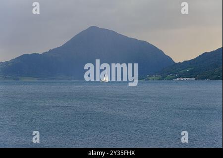 Una barca a vela solitaria si muove dolcemente su un tranquillo fiordo, incorniciato da montagne ondulate all'avvicinarsi del crepuscolo, creando un'atmosfera tranquilla. Foto Stock