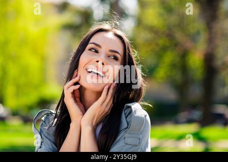 Foto ritratto di una bella giovane ragazza ispanica sorridente che indossa il cappotto grigio del parco locale della città che tocca le guance rilassate nella natura Foto Stock