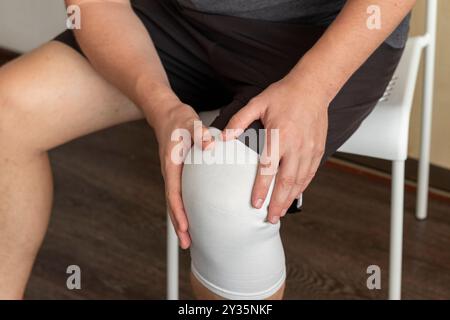 Colpo dell'uomo seduto sulla sedia bianca, che tiene il ginocchio con le mani, bendaggio elastico sul ginocchio destro Foto Stock