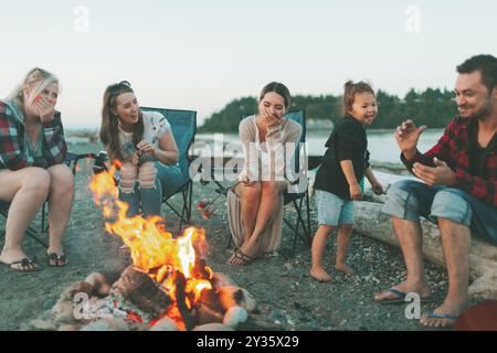 Un gruppo di amici seduti intorno a un falò sulla spiaggia al tramonto. Foto Stock