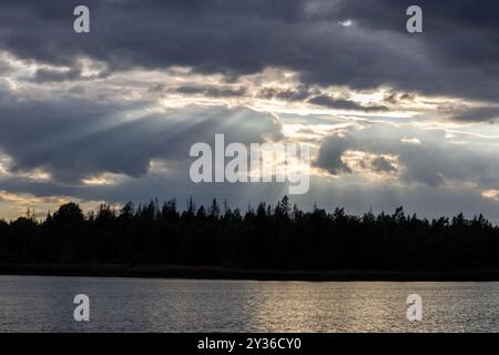 Un tranquillo paesaggio caratterizzato da un fiume calmo che riflette il cielo. Le nuvole scure si stagliano in alto, con raggi di luce solare che irradiano, illuminando il dist Foto Stock