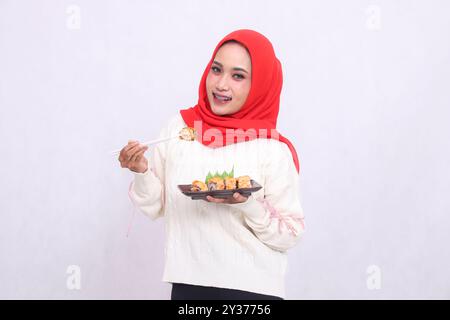 La ragazza asiatica che indossa un hijab è allegra per la macchina fotografica che prende le bacchette e porta con sé un piatto contenente sushi (cibo giapponese). Bella musulmana Wo Foto Stock