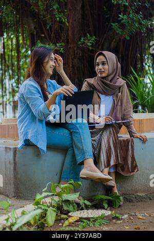 Un paio di allegre donne indonesiane schiette che discutono di studiare insieme trasportando laptop, libri e appunti nel parco del campus. studentesse per Foto Stock