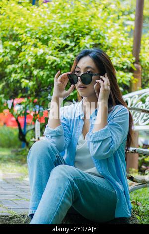 la donna indonesiana che indossa gli occhiali da sole è sorpresa di chiamare qualcuno tramite un dispositivo cellulare seduto sull'erba in un parco all'aperto. studentesse per la Foto Stock