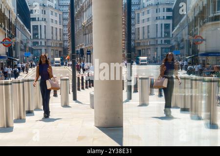 Riflesso nella finestra di una donna che cammina nella City di Londra con strade cittadine, edifici alti, pedoni e dissuasori fissi della City of London. Foto Stock