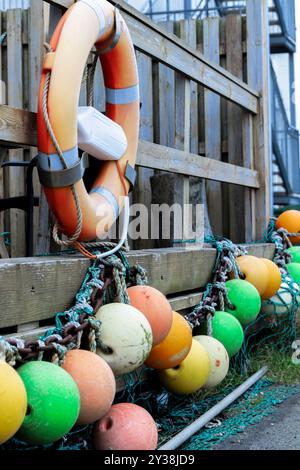 Una fila di boe arancioni e gialle sono legate ad una recinzione di legno, vicino ad un salvagente arancione Foto Stock