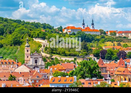 Il monastero di Strahov si trova in cima a una collina che domina la città di Praga. I tetti arancioni e le torri gemelle si stagliano contro la lussureggiante vegetazione della collina circostante. Sotto, una rete di tetti di tegole rosse si estende verso l'orizzonte, formando un arazzo vibrante e intricato. Il cielo sopra è una tela blu brillante, punteggiata da soffici nuvole bianche. Foto Stock