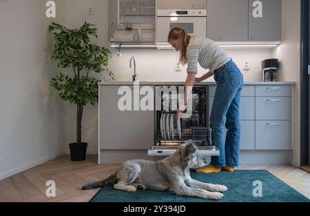 Donna che carica piatti sporchi in lavastoviglie e il suo cane sdraiato sul pavimento Foto Stock