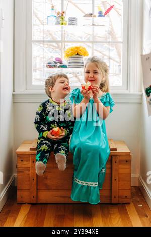 Due bambini seduti accanto a una finestra, mangiando felici mele Foto Stock