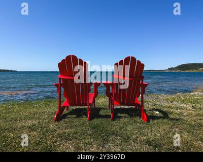 Due sedie rosse adirondack sull'erba che si affacciano sull'oceano nelle giornate di sole. Foto Stock