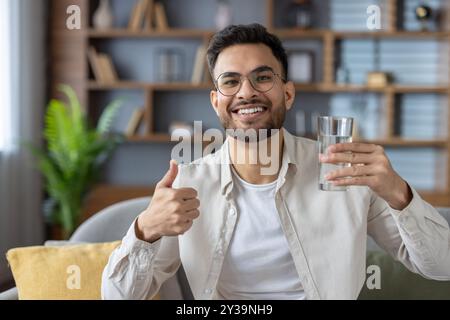 Uomo sorridente con bicchieri che reggono un bicchiere d'acqua che mostra i pollici in su. Si siede comodamente nel salotto moderno trasmettendo positività, salute e idratazione. Foto Stock