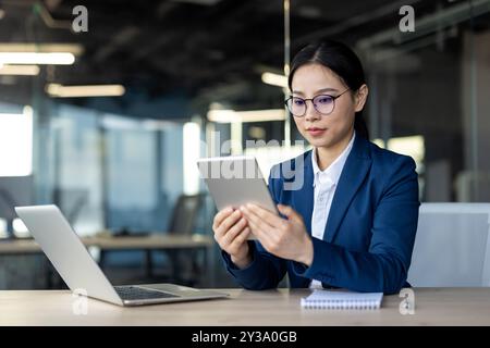 Donna d'affari asiatica si impegna con un tablet in ufficio. Scene mette in evidenza l'uso della tecnologia sul luogo di lavoro, con notebook e notebook aperti nelle vicinanze, sottolineando la produttività e le pratiche aziendali moderne Foto Stock