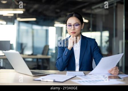 Donna d'affari asiatica che esamina documenti alla scrivania con grande serietà. Si concentra sull'analisi della documentazione e sulla definizione di strategie per le attività lavorative, sulla concentrazione e sul business professionale Foto Stock