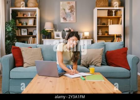 Donna sorridente che lavora in multitasking a casa, scrivendo appunti durante una riunione virtuale Foto Stock