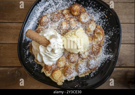 Waffle belgi con gelato alla vaniglia e panna montata, cialde, serviti su un piatto nero e spolverati con zucchero a velo, su un tavolo di legno, via superiore Foto Stock