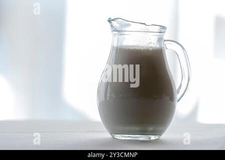 Caraffa di cristallo illuminata dal sole ripiena di latte, isolata su sfondo bianco. Foto Stock