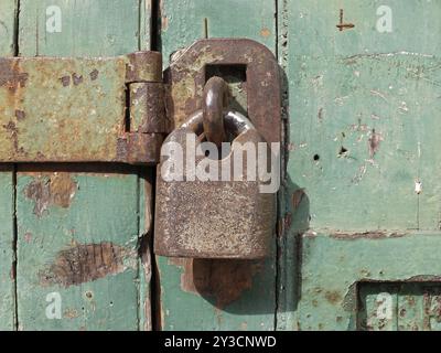 Primo piano di un vecchio lucchetto in metallo arrugginito che fissa una barra di ferro su una porta di legno verde Foto Stock