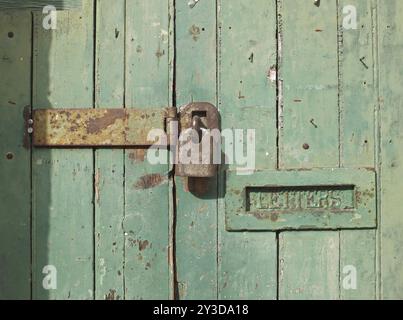 Primo piano di una vecchia porta di legno con vernice verde sbiadita, un lucchetto chiuso arrugginito e una vecchia cassetta delle lettere in metallo Foto Stock