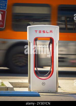 LOS ANGELES, CA - 25 giugno 2024: Stazione di ricarica Tesla Supercharger di fronte a una strada trafficata con l'autobus LA Metro in accelerazione sullo sfondo, motion BL Foto Stock