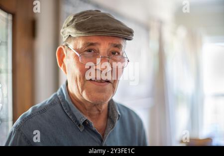 Ritratto di un uomo anziano sorridente e felice che indossa un berretto piatto e occhiali alla moda a casa, primo piano. DOF poco profondo, copyspace. Foto Stock
