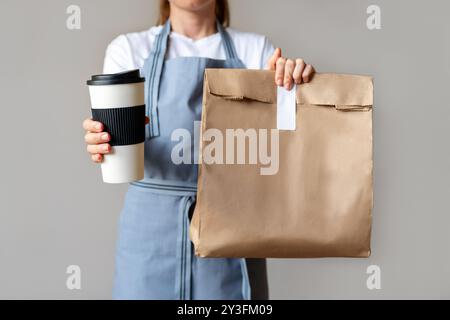 Cibo da asporto e bevande. Una donna in grembiule blu tiene in mano una tazza da caffè da viaggio e un sacchetto di carta riciclata con cibo Foto Stock