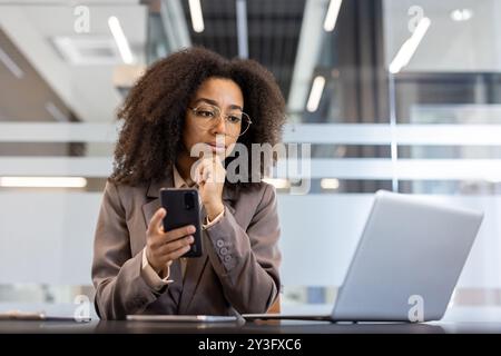 Giovane donna d'affari afroamericana seria e premurosa seduta in ufficio alla scrivania, tenendo il telefono in mano e guardando lo schermo del laptop. Foto Stock