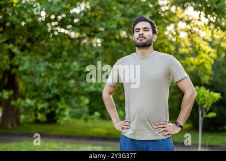 Sportivo sicuro in piedi in un parco, mani sui fianchi e momenti di relax. Cattura l'essenza dell'esercizio all'aperto, della routine fitness, della consapevolezza e della tranquillità in un ambiente naturale Foto Stock