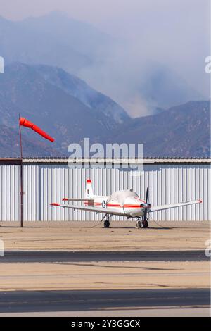 Un aereo privato ad ala bassa è legato vicino a un indicatore del vento, il che indica un'attività del vento moderata. Il fumo degli incendi boschivi è visibile su una montagna vicina sullo sfondo. Foto Stock