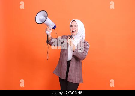 Giovane donna asiatica in hijab arrabbiata che guarda a destra schietta schietta schietta schietta con il microfono che tiene l'altoparlante a megafono isolato su sfondo arancione, Foto Stock