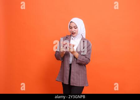 Il fascino di una bella donna asiatica che indossava l'hijab ha scioccato sia le mani tenendo e contando banconote di rupia isolate su sfondo arancione. finanza, stile di vita Foto Stock