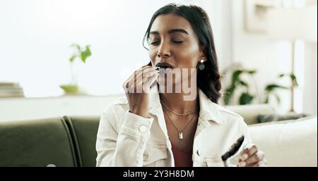 Cioccolatini, rilassati e sorridi con una donna indiana sul divano nel soggiorno dell'appartamento per una pausa o un riposo. Caramelle, cibo e dolci con una persona felice che mangia Foto Stock