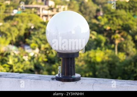 Lampada rotonda bianca in vetro su asta in acciaio nero montata sulla parete della casa, sfondo naturale sfocato. Foto Stock