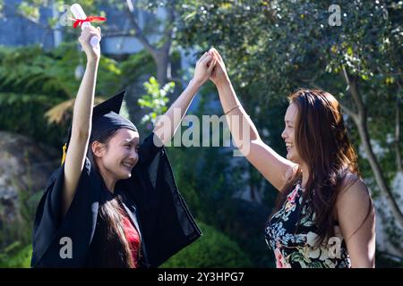 In giardino, celebrando la laurea, figlia asiatica in cappello e abito che tiene il diploma con la madre Foto Stock