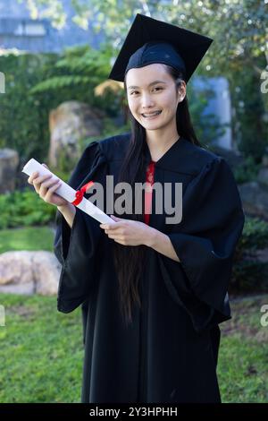 In giardino, si diploma di giovane donna asiatica in cappello e abito con diploma, sorridendo all'aperto Foto Stock