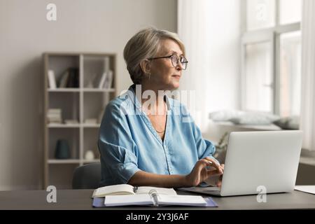 Una donna siede alla scrivania con un notebook e guarda con attenzione fuori dalla finestra Foto Stock