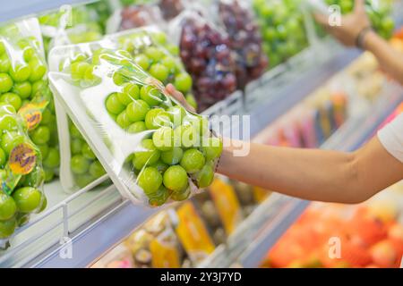 Una persona che cerca un pacco di uva verde in un negozio di alimentari. Gli scaffali sono riforniti di vari frutti, tra cui uve rosse e altri pacchetti Foto Stock